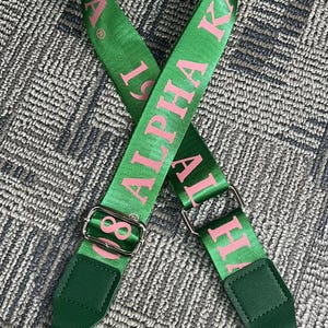 Puede incluir: Correa de bolso verde con texto rosa "Alpha Kappa Alpha". La correa presenta letras griegas rosas, herrajes met&aacute;licos plateados y detalles de polipiel verde. Dise&ntilde;ada para sujetar a un bolso.