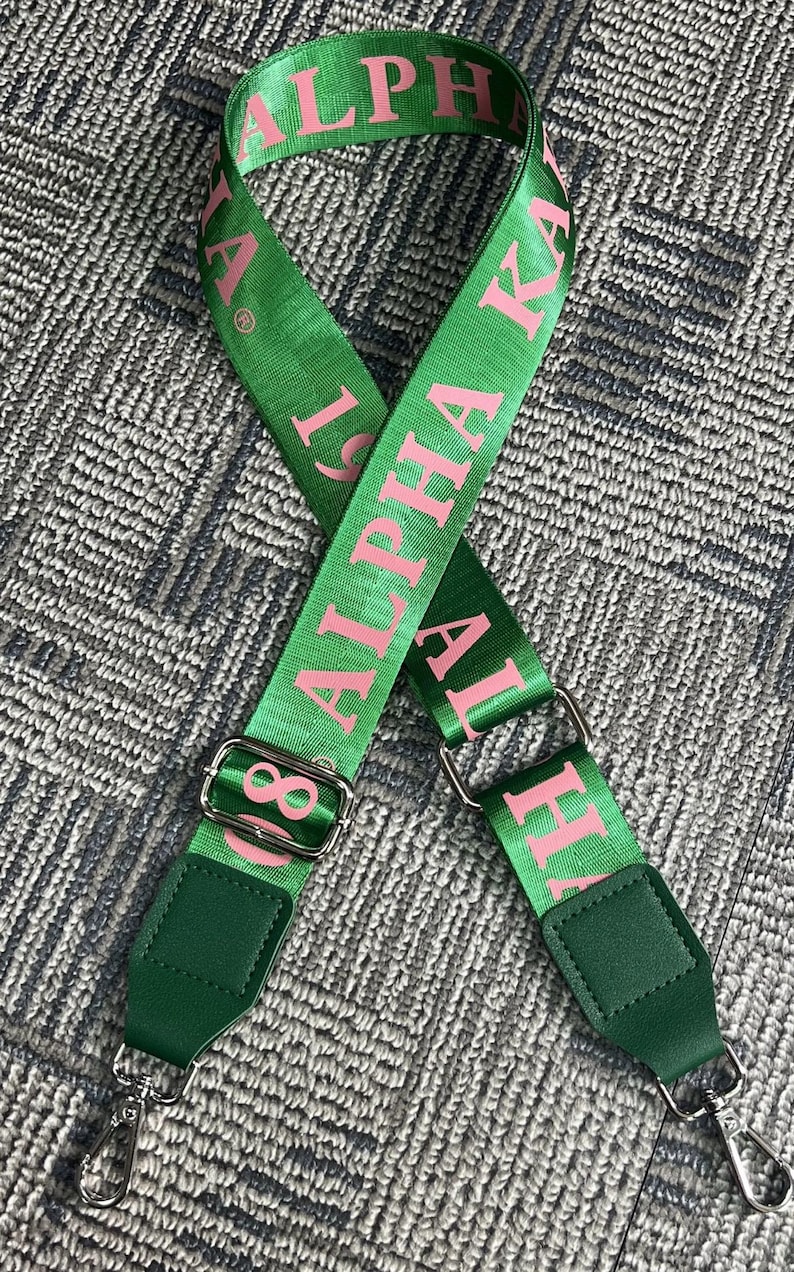 Puede incluir: Correa de bolso verde con texto rosa "Alpha Kappa Alpha". La correa presenta letras griegas rosas, herrajes met&aacute;licos plateados y detalles de polipiel verde. Dise&ntilde;ada para sujetar a un bolso.