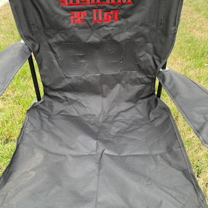 May include: Black folding outdoor chair with red embroidered text that reads "Michelle Fall '22" and "Gree". The chair is set up on a grassy surface.