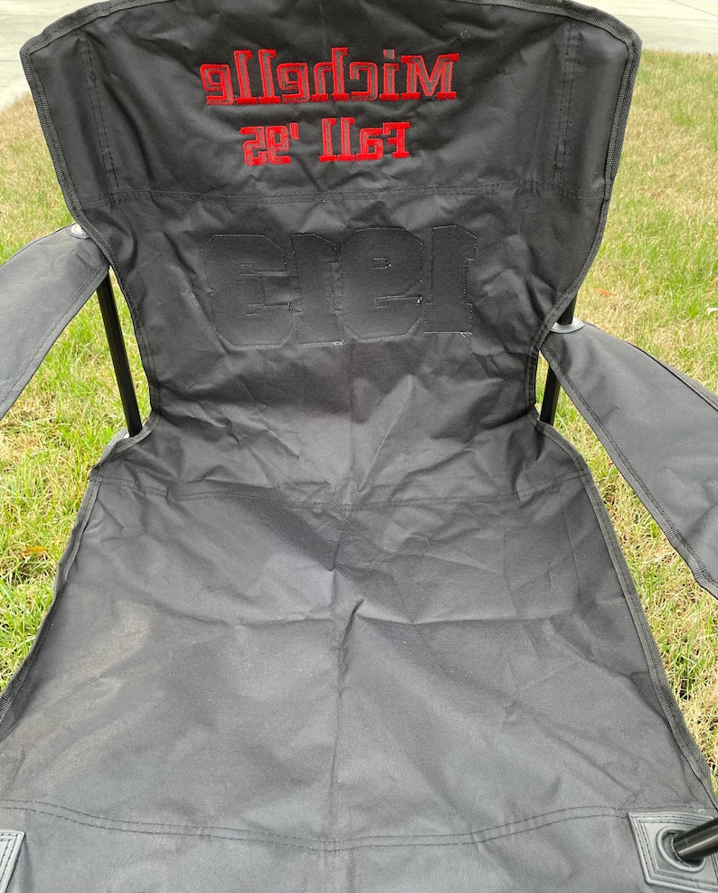 May include: Black folding outdoor chair with red embroidered text that reads "Michelle Fall '22" and "Gree". The chair is set up on a grassy surface.