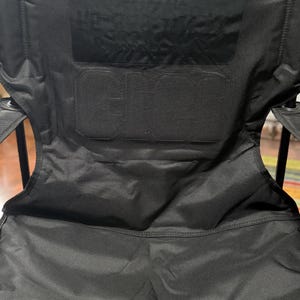 May include: Black folding camping chair with the text "GCI" and "OUTDOOR" embossed on the backrest. The chair has a sturdy frame and a durable fabric seat, perfect for outdoor activities.