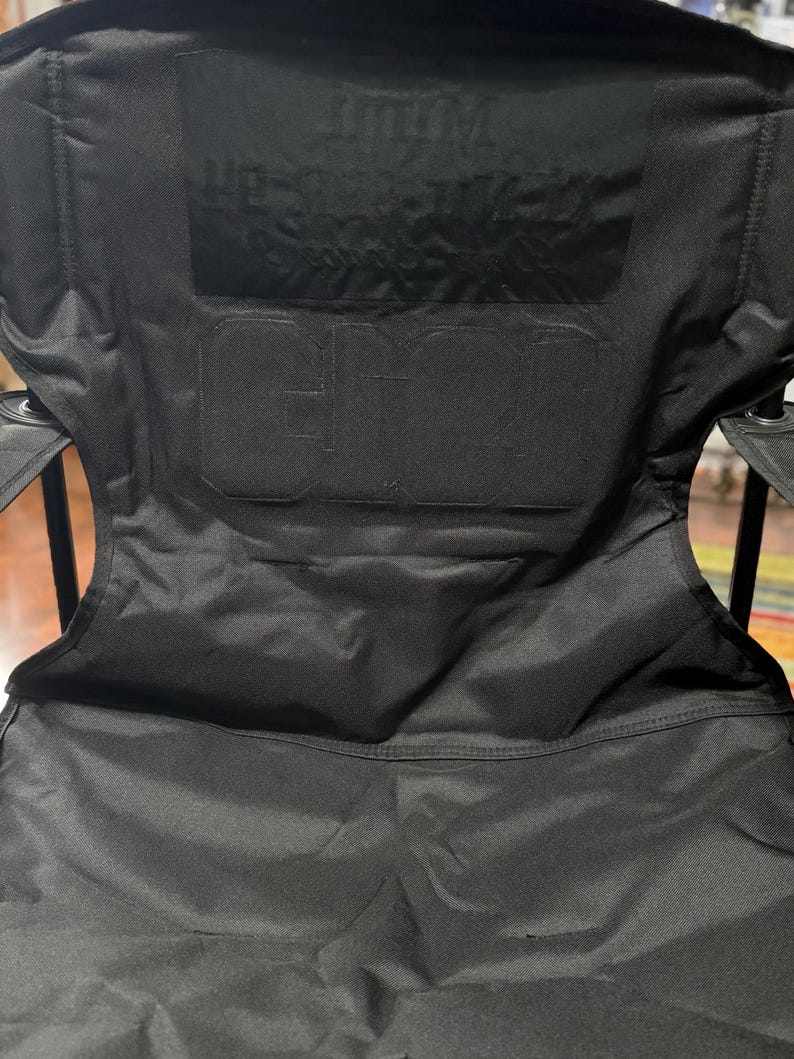 May include: Black folding camping chair with the text "GCI" and "OUTDOOR" embossed on the backrest. The chair has a sturdy frame and a durable fabric seat, perfect for outdoor activities.