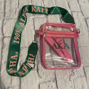 Puede incluir: Un bolso cruzado transparente con ribete y correa rosas. El bolso tiene una correa verde con el texto "ALPHA KAPPA ALPHA 1908". El bolso tambi&eacute;n tiene una correa rosa con las letras "AKA".