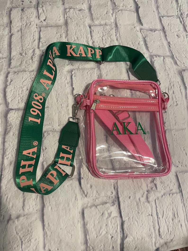 Puede incluir: Un bolso cruzado transparente con ribete y correa rosas. El bolso tiene una correa verde con el texto "ALPHA KAPPA ALPHA 1908". El bolso tambi&eacute;n tiene una correa rosa con las letras "AKA".