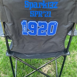 May include: Black folding chair with blue embroidered text that reads "Spark13Z SPR'21" and "1920". The chair is set up on a grassy lawn. The chair has a black frame and a black seat and back.