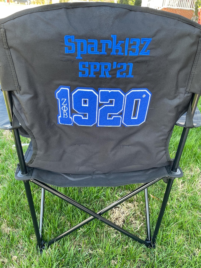 May include: Black folding chair with blue embroidered text that reads "Spark13Z SPR'21" and "1920". The chair is set up on a grassy lawn. The chair has a black frame and a black seat and back.
