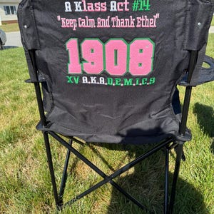 May include: Black folding chair with a black frame, set on green grass. The chair back features pink and green text that reads "A Klass Act #14", "Keep Calm And Thank Ethel", "1908", and "XV A.K.A.D.E.M.I.C.S".
