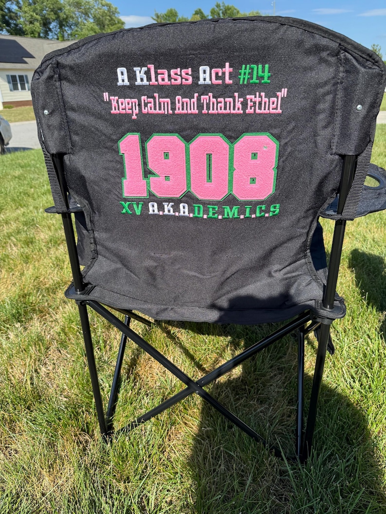 May include: Black folding chair with a black frame, set on green grass. The chair back features pink and green text that reads "A Klass Act #14", "Keep Calm And Thank Ethel", "1908", and "XV A.K.A.D.E.M.I.C.S".