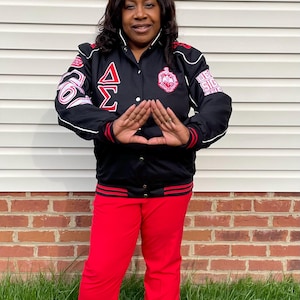 May include: A woman wearing a black and red varsity jacket with the letters "Delta Sigma Theta" embroidered on the front. She is making the sorority's signature hand sign.