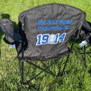 May include: Black folding chair with the text "Phi Beta Sigma Fraternity Inc 1914" embroidered in blue and white. The chair has a black frame and armrests, and a small zippered pocket on the side.