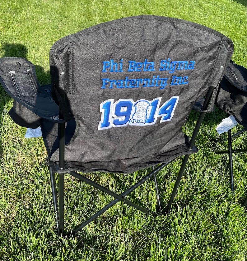 May include: Black folding chair with the text "Phi Beta Sigma Fraternity Inc 1914" embroidered in blue and white. The chair has a black frame and armrests, and a small zippered pocket on the side.
