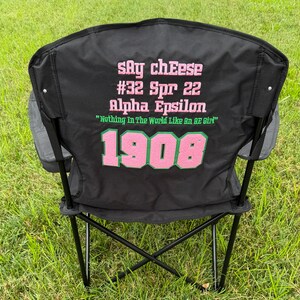 May include: Black folding chair with text in pink and green. The text includes "Say cheese", "#32 Spr 22", "Alpha Epsilon", "Nothing In The World Like An AE Girl", and "1908". The chair has gray side pockets and is set on a grassy surface.