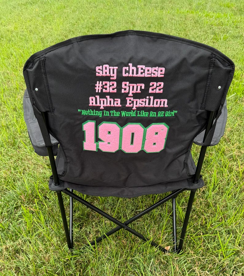 May include: Black folding chair with text in pink and green. The text includes "Say cheese", "#32 Spr 22", "Alpha Epsilon", "Nothing In The World Like An AE Girl", and "1908". The chair has gray side pockets and is set on a grassy surface.