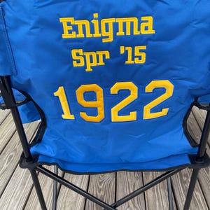 May include: Blue folding chair with black metal frame. The chair back features the text "Enigma Spr '15 1922" in yellow block letters. The chair is set up on a wooden deck.