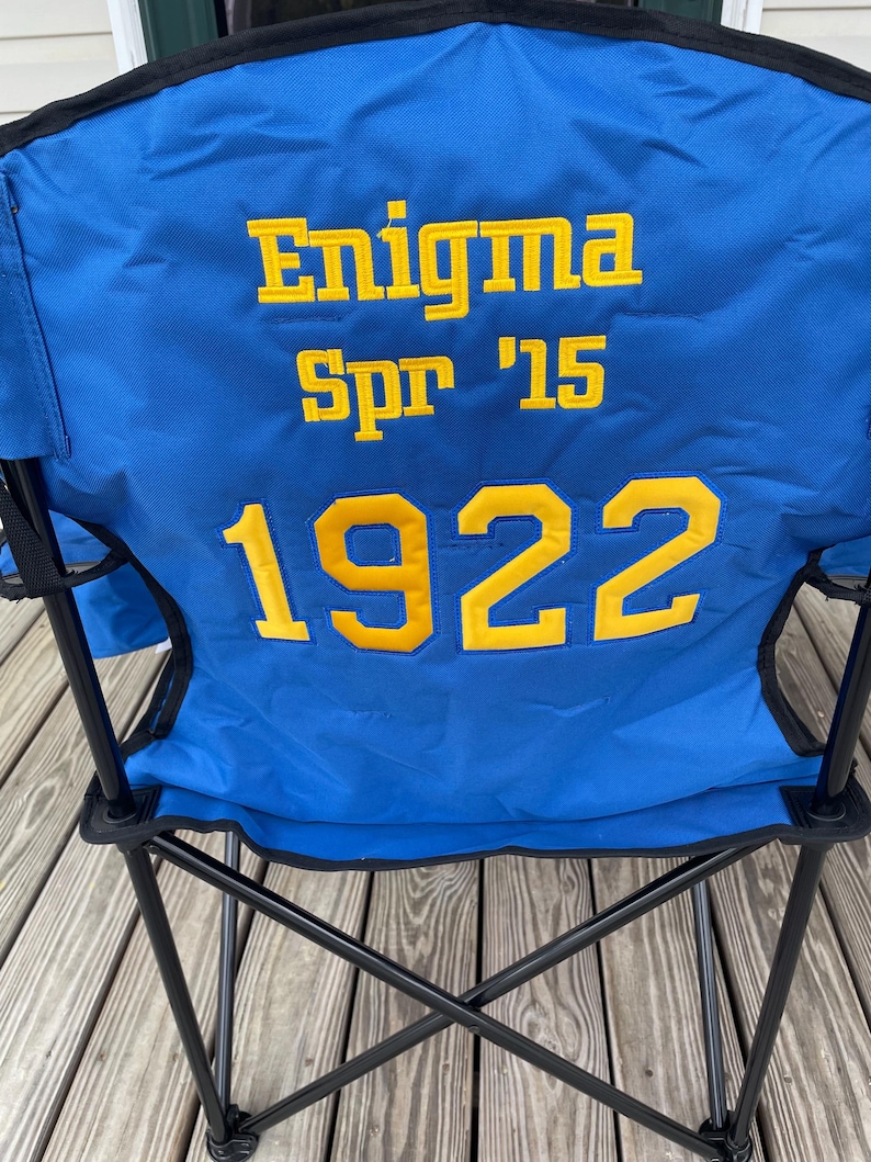 May include: Blue folding chair with black metal frame. The chair back features the text "Enigma Spr '15 1922" in yellow block letters. The chair is set up on a wooden deck.