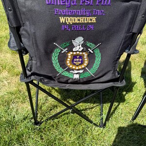 May include: Black folding chair with the text "Omega Psi Phi Fraternity, Inc. WOODCHUCK #4, FALL 24" in purple and gold. The chair features a crest with swords and a laurel wreath in green, gold, and purple.