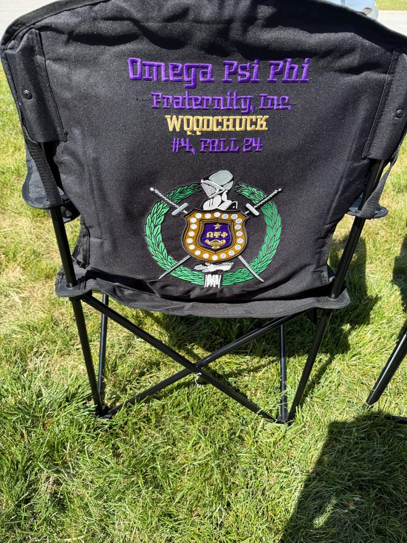 May include: Black folding chair with the text "Omega Psi Phi Fraternity, Inc. WOODCHUCK #4, FALL 24" in purple and gold. The chair features a crest with swords and a laurel wreath in green, gold, and purple.