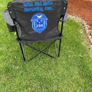 May include: Black folding outdoor chair with the text "Zeta Phi Beta Sorority, Inc." in blue. The chair back features a blue and white emblem with the letters "ZFB" and the founding year, 1920.
