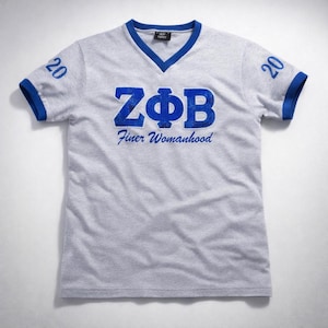 May include: A gray v-neck t-shirt with blue trim on the collar and sleeves. The shirt features the Greek letters ZΦB in blue, with the words "Finer Womanhood" below. The number "20" is printed on each sleeve.