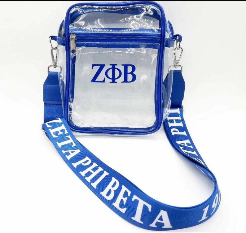 Puede incluir: Bolso transparente de pl&aacute;stico con cierre de cremallera azul y correa azul. La correa tiene las palabras "Zeta Phi Beta" impresas en blanco. El bolso tiene una cremallera azul y las letras "Z&Phi;B" impresas en azul.