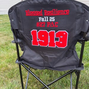 May include: Black folding chair with the words "Blessed Resilience" and "Fall 25 #27 PAC" in red lettering. The number "1913" is also displayed in red with a white outline. The chair has a black metal frame and is set on a grassy lawn.