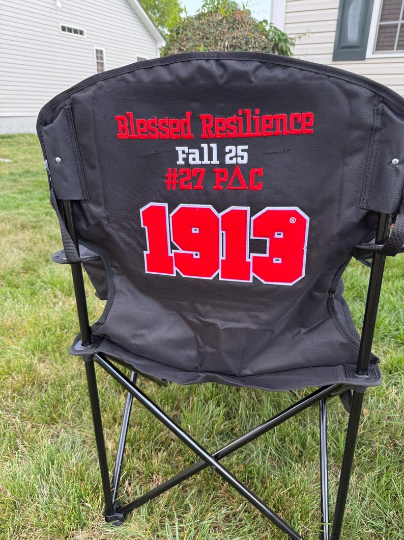 May include: Black folding chair with the words "Blessed Resilience" and "Fall 25 #27 PAC" in red lettering. The number "1913" is also displayed in red with a white outline. The chair has a black metal frame and is set on a grassy lawn.