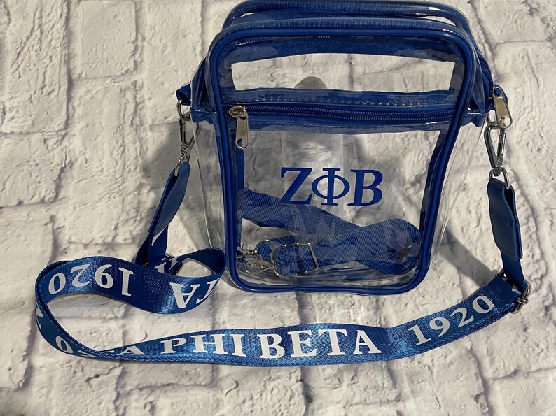 Puede incluir: Un bolso de hombro transparente azul y blanco con una correa azul. La correa tiene el texto "Z&Phi;B" y "Alpha Phi Beta 1920" impreso en ella.