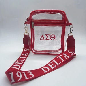 May include: Clear plastic crossbody bag with a red trim and a red adjustable strap. The strap has the text "1913 DELTA SIGMA" printed in white. The bag has a red zipper and the Greek letters "ΔΣΘ" printed in red on the front.