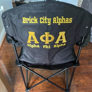 May include: Black folding chair with the text "Brick City Alphas" and "A&Phi;A Alpha Phi Alpha" in gold lettering. The chair has a black frame and a black fabric seat and back. The chair is designed for outdoor use.