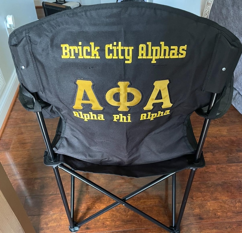 May include: Black folding chair with the text "Brick City Alphas" and "A&Phi;A Alpha Phi Alpha" in gold lettering. The chair has a black frame and a black fabric seat and back. The chair is designed for outdoor use.