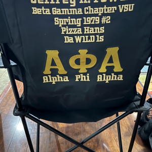 May include: Black folding chair with gold lettering. The text on the chair reads "Jeffrey H. Powell, Beta Gamma Chapter VSU, Spring 1979 #2, Pizza Hans, Da WILD Is, Alpha Phi Alpha". The chair is set up on a wooden floor.