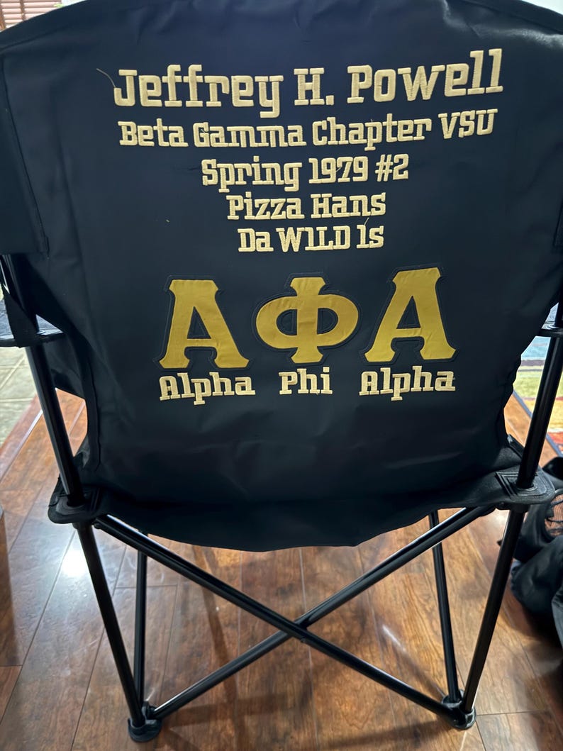 May include: Black folding chair with gold lettering. The text on the chair reads "Jeffrey H. Powell, Beta Gamma Chapter VSU, Spring 1979 #2, Pizza Hans, Da WILD Is, Alpha Phi Alpha". The chair is set up on a wooden floor.