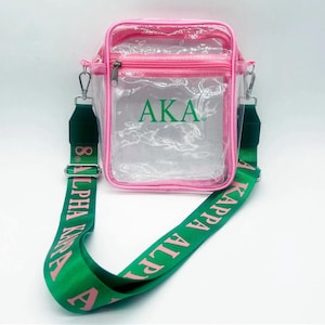 May include: A clear crossbody bag with a pink trim and a green strap. The bag features the letters "AKA" in green on the front. The green strap has the words "ALPHA KAPPA ALPHA" printed in pink.
