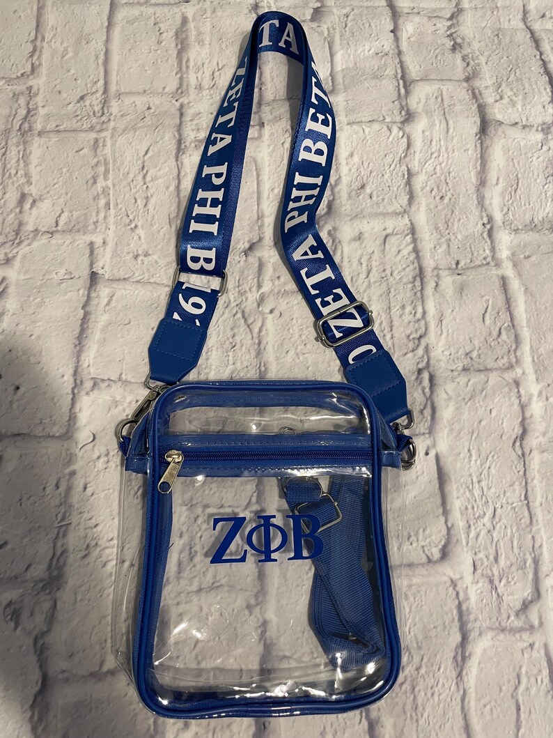 Puede incluir: Una bolsa de bandolera transparente de pl&aacute;stico con ribete azul y una correa ajustable azul. La correa tiene el texto "Zeta Phi Beta 19/" impreso en ella. La bolsa tiene una cremallera azul y las letras "Z&Phi;B" est&aacute;n impresas en la parte delantera de la bolsa.
