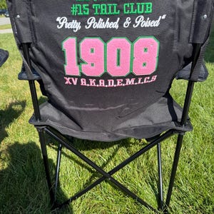 May include: Black folding chair with the text "KOVER GIRL #15 TAIL CLUB" in green and white. The text "Pretty, Polished, & Poised" is also on the chair. The number "1908" is in pink and green, with "XV A.K.A.D.E.M.I.C.S" below.