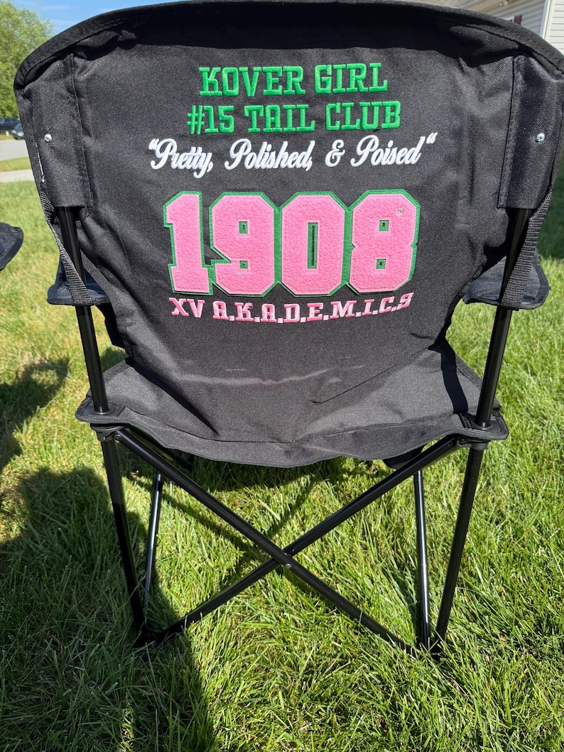 May include: Black folding chair with the text "KOVER GIRL #15 TAIL CLUB" in green and white. The text "Pretty, Polished, & Poised" is also on the chair. The number "1908" is in pink and green, with "XV A.K.A.D.E.M.I.C.S" below.