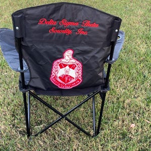 May include: Black folding chair with the words "Delta Sigma Theta Sorority, Inc" in red script. The chair has a red and white emblem on the back. The chair is set up on a grassy surface.