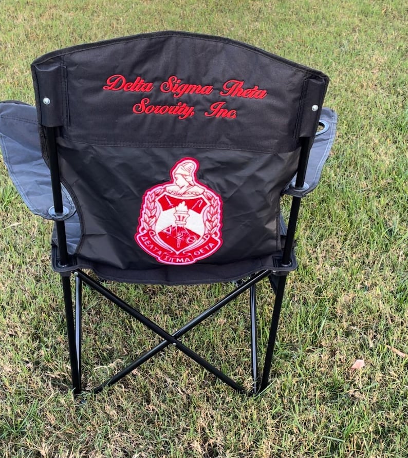 May include: Black folding chair with the words "Delta Sigma Theta Sorority, Inc" in red script. The chair has a red and white emblem on the back. The chair is set up on a grassy surface.