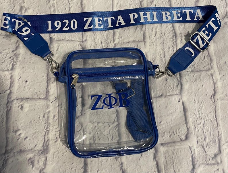 Puede incluir: Un bolso transparente de pl&aacute;stico con un ribete azul y una correa azul. La correa tiene el texto "1920 ZETA PHI BETA" impreso en blanco. El bolso tiene las letras "Z&Phi;B" impresas en azul en la parte delantera.