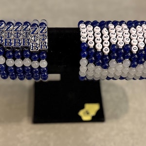 Puede incluir: Dos pulseras de cuentas expuestas en un soporte negro. Una pulsera tiene cuentas azules y blancas con dijes de letras plateadas que deletrean "Z B" y "Ф". La otra pulsera tiene cuentas blancas con los números "1920" y cuentas azules.