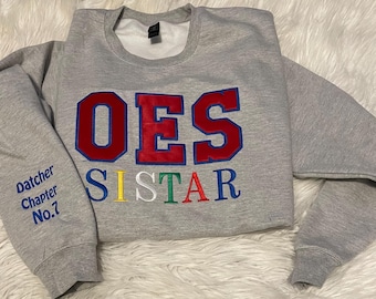 May include: A gray sweatshirt with the letters "OES SISTER" in red, blue, yellow, and green embroidery. The sleeve has "Datcher Chapter No. 7" embroidered in blue. The sweatshirt is on a white fluffy surface.