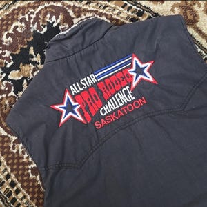 May include: A dark grey vest featuring the embroidered text "ALL STAR PRO RODEO CHALLENGE SASKATOON" in red, white, and blue. The design includes two star shapes. The vest has a western style.