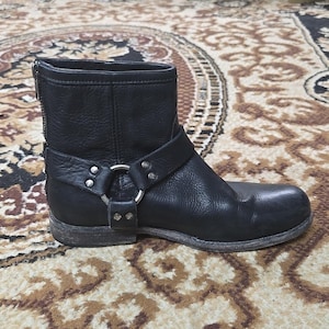May include: Black leather ankle boot with a harness strap and silver-tone hardware. The boot has a zip closure at the back and a rounded toe. The boot is on a patterned rug.