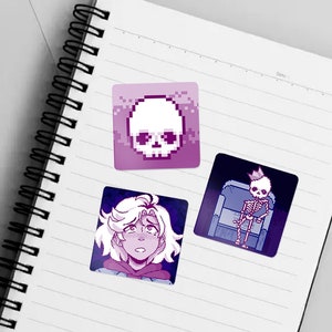 May include: Three pixel art stickers on a lined notebook. The first sticker is a purple and white skull. The second sticker is a purple and white character with short, wavy hair. The third sticker is a purple and white skeleton wearing a crown and sitting on a throne.
