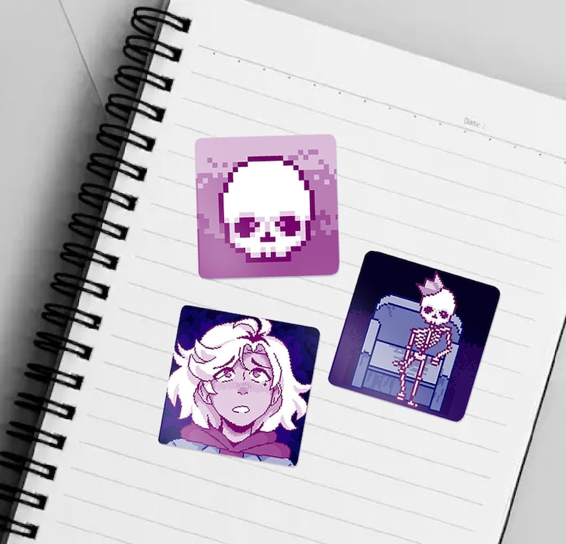 May include: Three pixel art stickers on a lined notebook. The first sticker is a purple and white skull. The second sticker is a purple and white character with short, wavy hair. The third sticker is a purple and white skeleton wearing a crown and sitting on a throne.