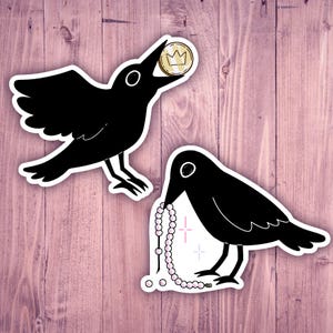 Puede incluir: Dos pegatinas de pájaros negros. Uno sostiene una moneda de oro con una corona, el otro una cadena de cuentas rosas. Ambos pájaros tienen contornos blancos sobre un fondo de madera rosa.