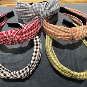 May include: Five fabric headbands with knotted tops. The headbands are in various colours: red, black and white, light blue, pink, and green. Each headband has a gingham pattern. The headbands are on a blue surface.