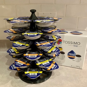 May include: A black plastic coffee pod holder with a circular base and multiple tiers. The holder is filled with Tassimo coffee pods, featuring a blue and yellow design with the text "Maxwell House" and "Tassimo". A box of Tassimo Maxwell House coffee pods is visible in the background.
