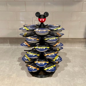 May include: A black and red Mickey Mouse head figurine sits atop a black metal coffee pod holder. The holder is filled with coffee pods from Maxwell House and Tassimo.