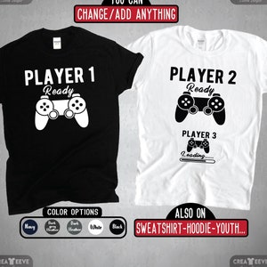 May include: Two t-shirts with a video game theme. The black t-shirt says "Player 1 Ready" with a white video game controller. The white t-shirt says "Player 2 Ready" with a black video game controller. The white t-shirt also says "Player 3 Loading..." with a black video game controller. The t-shirts are available in navy, dark heather, white, and black.